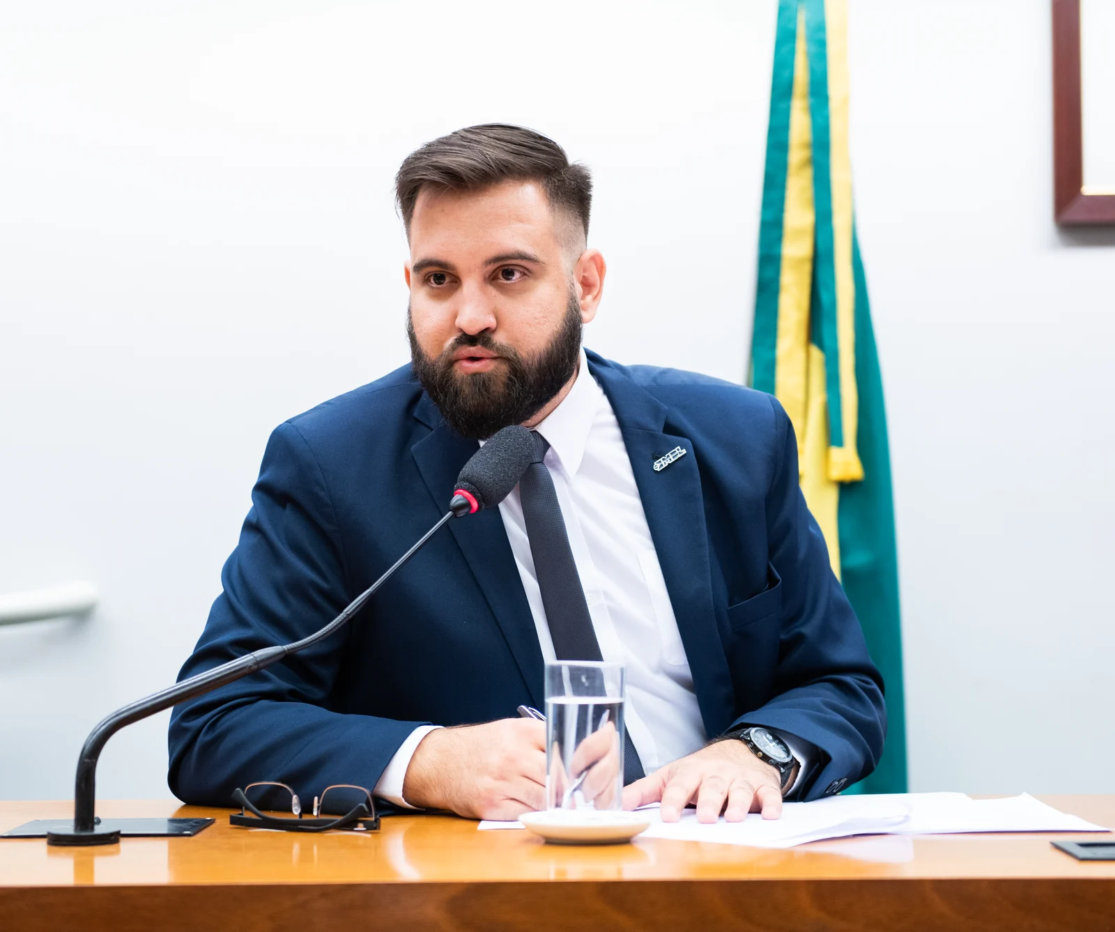 Warlen Miiller fala ao microfone em uma mesa da Câmara dos Deputados, com a bandeira do Brasil ao fundo